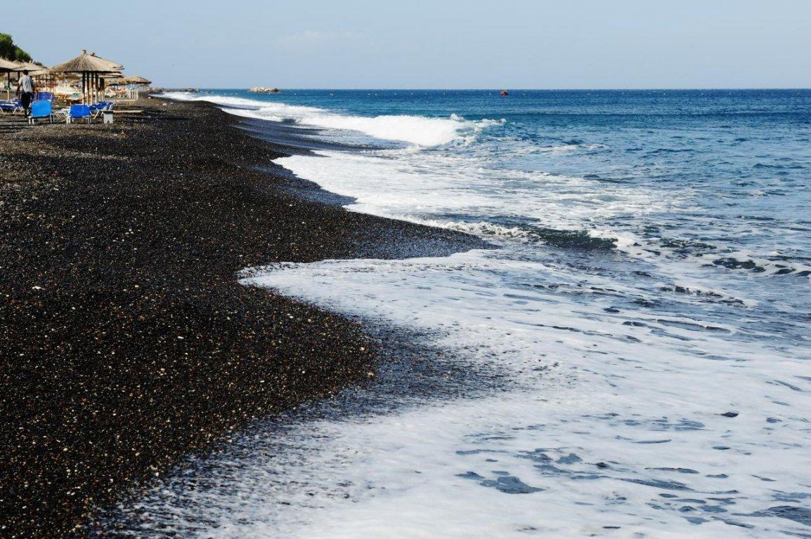 пляж Камари. На нем черный песок с галькой такого же цвета омывается изумительно чистым морем. За чистоту и экологичность этот пляж ежегодно получает «Голубой флаг».